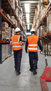 Two male warehouse employees in safety vests and hard hats walking through an aisle. One colleague is pulling a pallet jack and talking