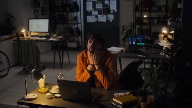 Caucasian young adult man working late in dark office, covering face in exhaustion while typing on laptop. Perfect for commercial use in videos about burnout, stress, and deadline pressure