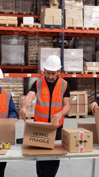 Mature warehouse worker in a safety vest and hard hat meticulously packing a cardboard box labeled vetro fragile in a distribution center