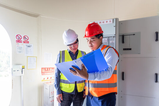 Two professional male workers in safety helmets and vests review documents with a clipboard inside an industrial indoor facility.