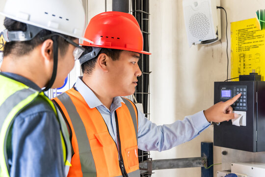 Two adult male workers in safety gear operate and inspect an industrial control device in an indoor workplace.
