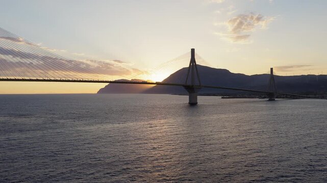 Rio-Antirrio Bridge with sunset over Gulf of Patras near Patra Greece, drone ascend tracking left reveals beautiful sun flare