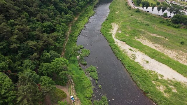 Aerial view follows shallow Pyeongchang River rocky bed and riverside trail beneath forested mountain near campsite