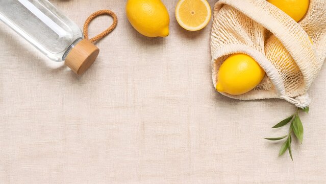 Flat lay of fresh organic lemons in a reusable mesh bag with a glass water bottle on a natural linen background, minimalist eco-friendly lifestyle concept with copy space for sustainable