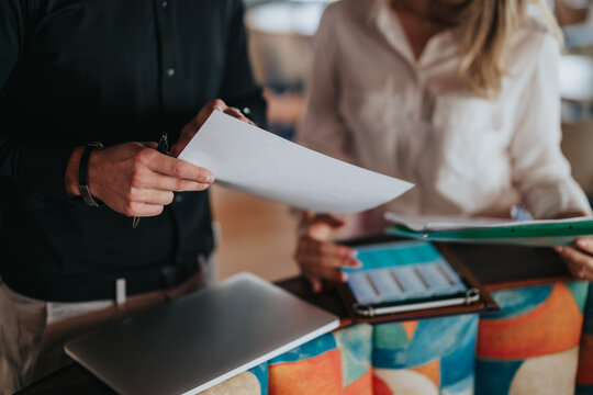 Two colleagues review printed documents and digital notes while standing at a workspace. They discuss files using a laptop and tablet during a casual remote work meeting.