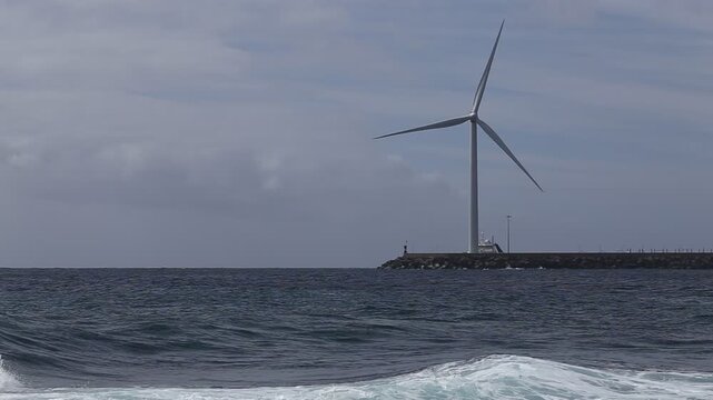 Canary Islands, Spain - March 12, 2016: Wind turbine at a harbour breakwater as a boat leaves a foamy wake across the Atlantic Ocean in a fixed HD shot