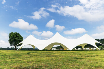 A large white tensile tent structure stands on a green grass field under a bright blue sky with scattered clouds, set in an open outdoor landscape. © tai