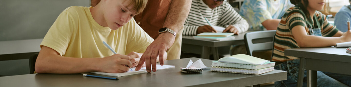 Header of teen students studying and writing in classroom while teacher guiding schoolwork, supporting learning and academic progress. Wide shot, ideal for education marketing and school promotion