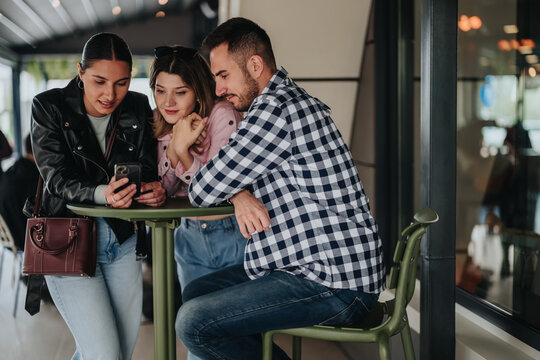 Three young friends gathered around a smartphone, smiling and focused on the screen. The casual group leans on a small cafe table while sharing content on the phone.
