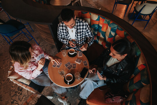 Three friends sit in a curved booth playing a card game while drinking coffee. The group enjoys a relaxed moment together around a small round table in a cafe.