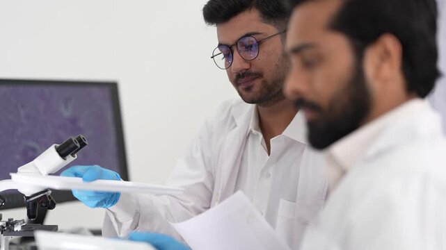 A bearded scientist in glasses holds up a stained histology tissue slide to show a colleague beside a microscope and monitor in a pathology lab.