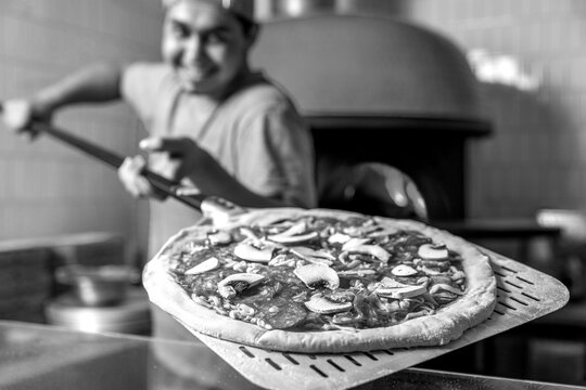 Chef placing the pizza in the hot oven to be baked. The chef prepares pizza with a shovel in his hands. Black and white