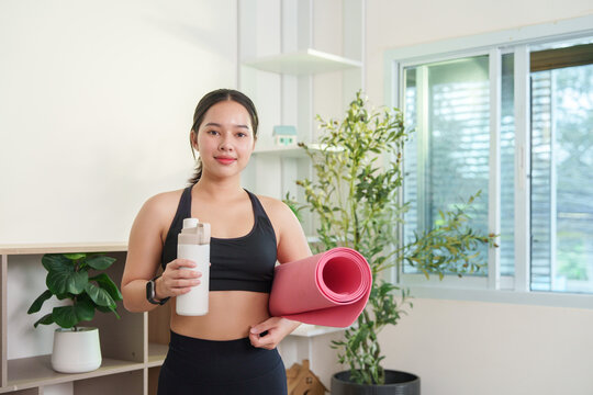 Asian woman holding yoga mat and water bottle for home workout