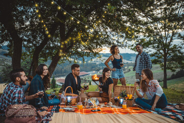 Obraz premium Group of young friends celebrating and sharing a happy outdoor picnic with wine, food, and music, enjoying togetherness in a scenic vineyard landscape during sunset