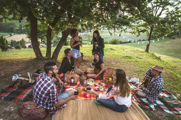Obraz premium Group of young friends celebrating and sharing a happy outdoor picnic with wine, food, and music, enjoying togetherness in a scenic vineyard landscape during sunset