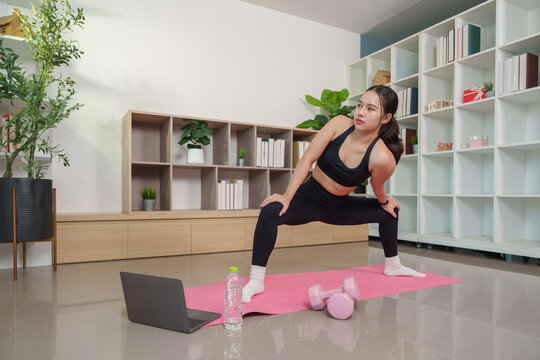 Asian woman stretching on mat with laptop and dumbbells for home workout