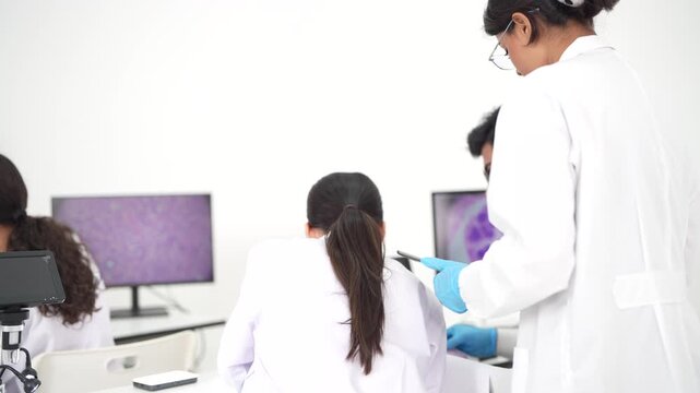 Research scientists work at a row of computer workstations displaying magnified cell tissue while a colleague reviews notes on a tablet.