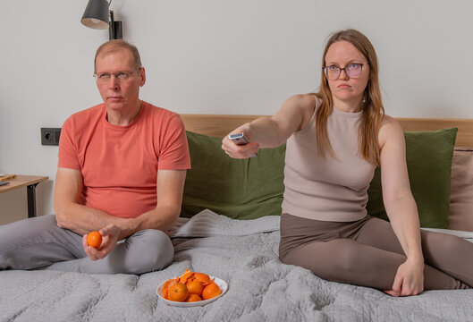 Woman switching channels while man eats tangerines. Couple relaxing on bed with snack and remote at