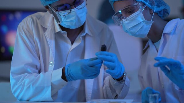 Under dramatic blue light, two masked scientists in caps closely handle a small sample at a bench with molecular graphics glowing behind.