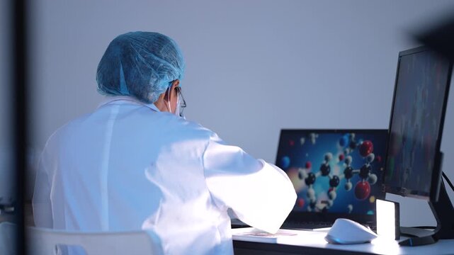 A capped scientist works at a microscope in a blue-lit lab as a molecular structure model rotates on the computer monitor.