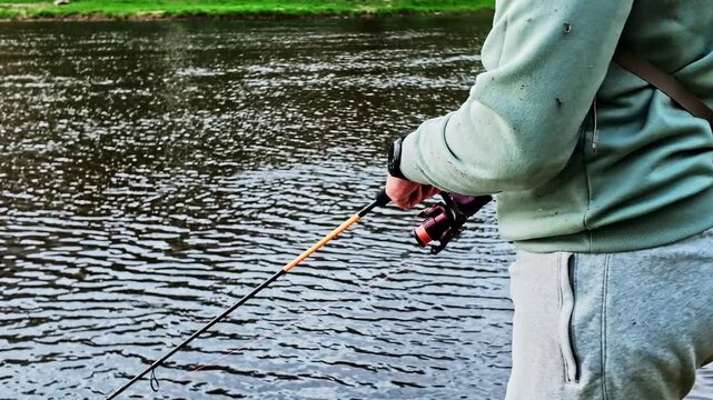 Close up on the hands of a fisherman spinning the reel of his fishing rod on a calm river