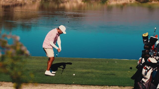 A golfer hits a low driving stinger across a blue water hazard toward the fairway at a desert course in slow motion