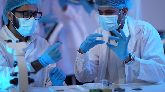 Two bearded scientists in masks carefully transfer a sample with a dropper over specimen dishes in a blue-lit research laboratory.