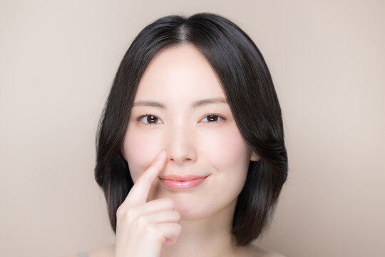 ピンクベージュ背景笑顔の美しい鼻のカメラ目線の日本人女性正面　美しい素肌とナチュラルメイクの美容クローズアップポートレート　小鼻を指を差す