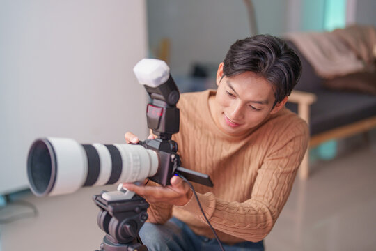 Asian male photographer adjusting professional camera with telephoto lens on tripod at home
