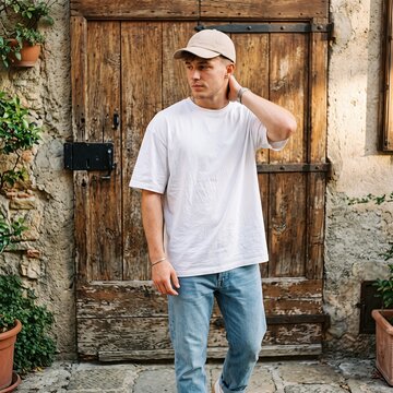 Young man wearing a white t-shirt and baseball cap stands in front of a rustic wooden door in an outdoor setting.