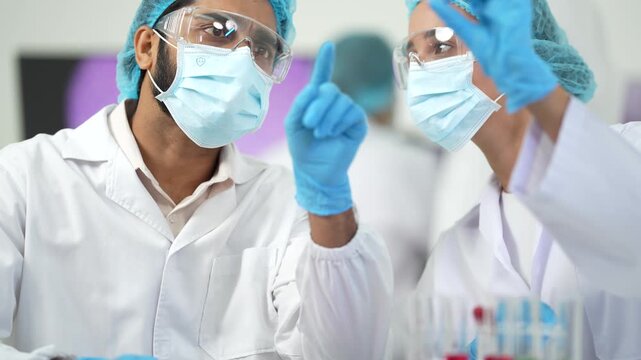 Two masked scientists hold up a red-liquid test tube to examine it closely above a rack of colourful samples.