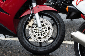 Obraz premium Close up view of front wheel of red sport motorcycle standing on wet asphalt road, showing intricate details of disc brake system, suspension, and rain droplets on fender