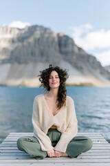 Fototapeta premium Young woman meditating on a dock by a mountain lake for serene wellness background and banner design