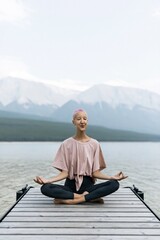 Fototapeta premium Smiling individual practicing seated meditation on a dock facing a mountain lake for mindfulness background and wellness banner with soft natural tones