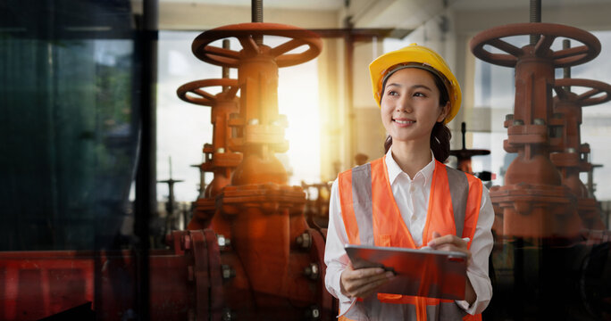 A smiling female engineer wearing a safety helmet and reflective vest holding a digital tablet in an industrial factory setting with large valves in the background.