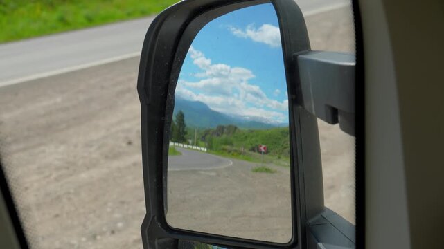 Point of view shot from a moving car showing a beautiful mountain landscape and blue sky with clouds reflecting in the side view mirror, embodying the concept of travel and adventure