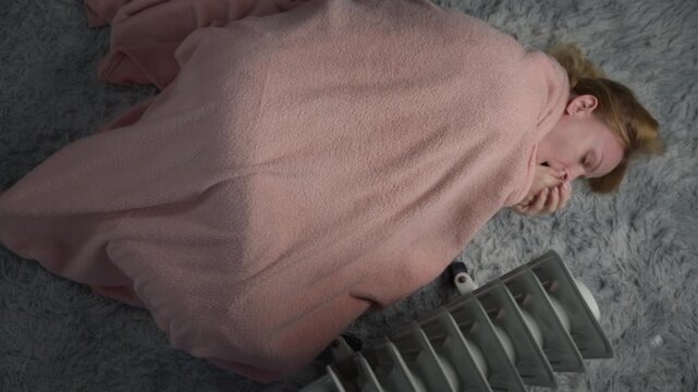 Young woman struggling to stay warm while sleeping beside an electric radiator at home. Rising heating costs, inflation, cost of living crisis and financial stress concept.