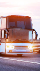 Front view of large modern passenger bus driving on highway at golden hour with bright headlights and warm sunlight lens flare, clear sky backdrop, vertical photo © HENADZY