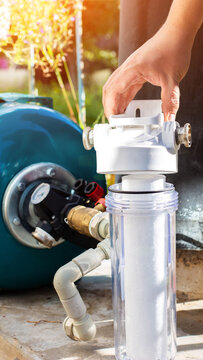 Hand of person installing or maintaining water filter system outdoors, clear filter housing, blue pressure tank, brass fittings, and pressure gauge under bright sunlight, vertical photo