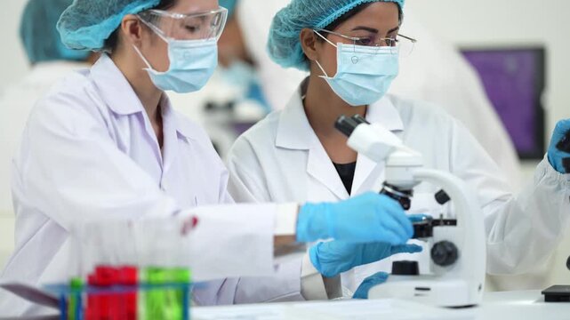A female scientist looks into a microscope while a colleague holds a test tube beside a rack of bright red samples.