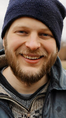 Obraz premium Close-up portrait smiling adult man with beard and blue knit cap looking at camera outdoors warm clothing natural light on face genuine expression vertical photo