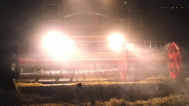 A combine harvester cuts barley at night as a dense cloud of dust rises around it. Bright headlights and a red cutting header give the rural harvest scene dramatic and cinematic appearance after dark.