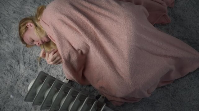 Young woman sleeping beside an electric radiator at home, highlighting the need for constant heating during cold weather and the rising cost of household energy consumption.