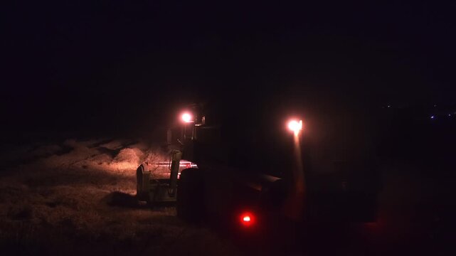 A combine harvester works through a ripe barley field at night moving under bright headlights. Illuminated straw rows, rising dust and the dark rural setting create a dramatic cinematic harvest scene.