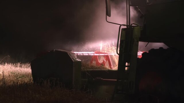 A combine harvester cuts barley at night as a dense cloud of dust rises around it. Bright headlights and a red cutting header give the rural harvest scene dramatic and cinematic appearance after dark.