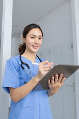 A smiling Asian female nurse or doctor wearing blue scrubs and a stethoscope, holding and using a...