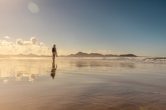A woman walking barefoot along the wet sandy shore of Famara beach in Lanzarote, Canary Islands, holding her sandals. Scenic coastal landscape with ocean reflections and vacation vibe.