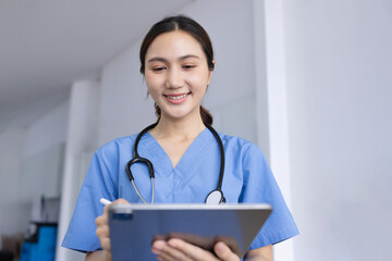 A smiling Asian female nurse or doctor wearing blue scrubs and a stethoscope, holding and using a...