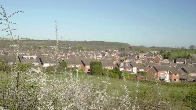 White-blossomed shrubs frame a grassy field leading to rows of suburban homes. Beyond them, open countryside and distant wind turbines complete the calm landscape.