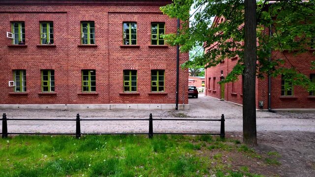 Historic red brick building in Ksiezy Mlyn district in Lodz Poland, green window frames and door, black metal fence, cobblestone walkway, green grass lawn, daylight, industrial heritage architecture.